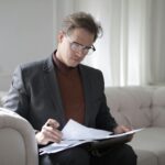 Elegant adult man in jacket and glasses looking through documents while sitting on white sofa in luxury room